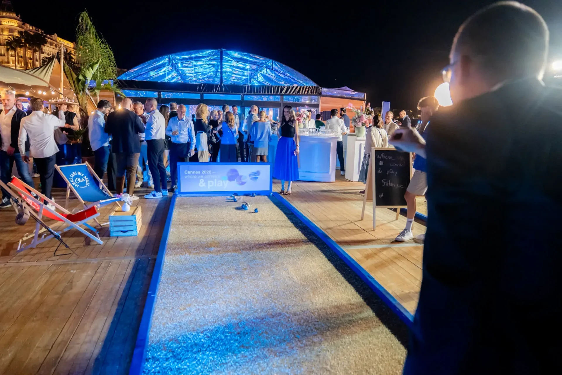 Piste de pétanque sur le deck de la plage, zone Connect & Play de la soirée de gala Heinemann à Cannes, avec la verrière illuminée en bleu au fond.