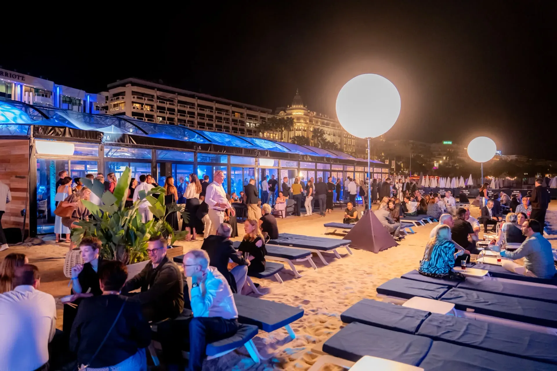 Invités détendus sur les transats et le sable de la plage de Cannes, espace Connect & Chill, la Croisette illuminée au fond, soirée de gala d'entreprise.
